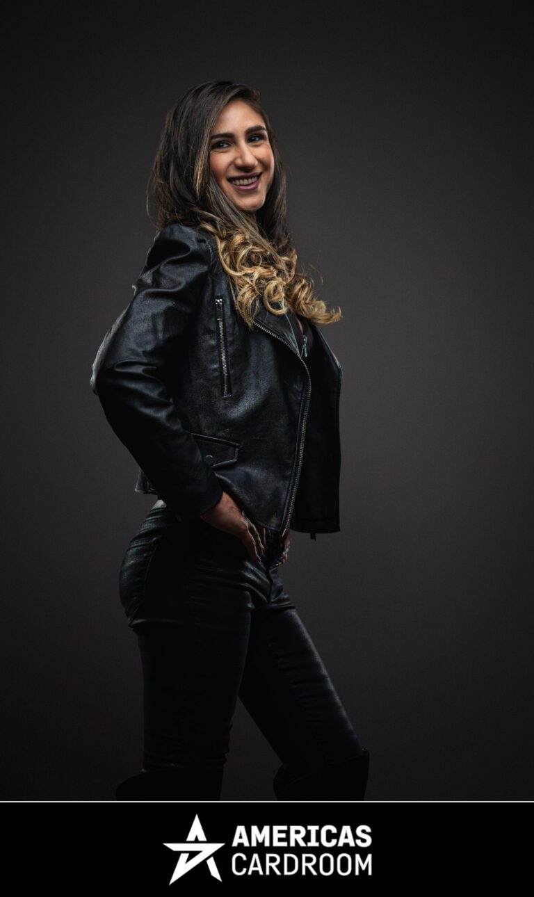 A three-quarter profile shot of Ana Marquez, an ACR poker pro, smiling and looking back towards the camera. She has long, dark hair with blonde highlights and is wearing a black leather motorcycle jacket and black shiny pants, with her hands on her hips, against a dark background.