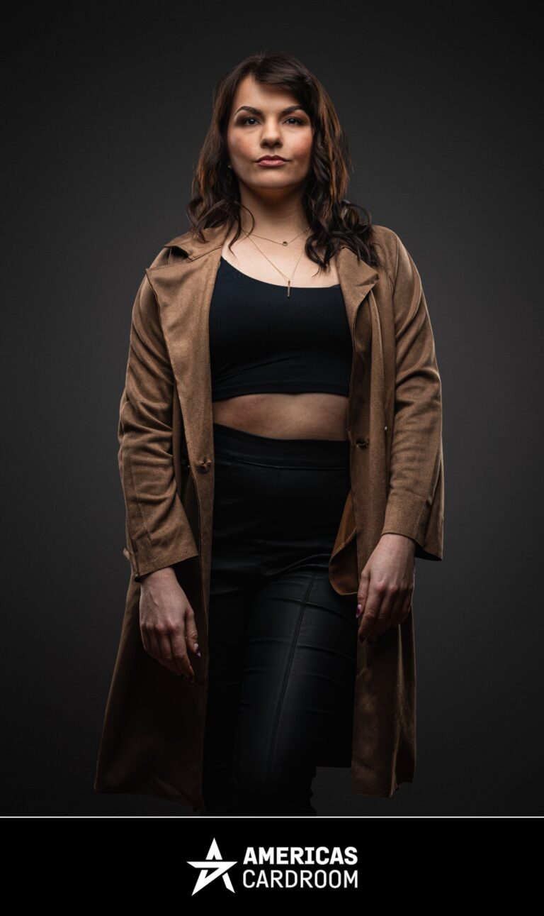 A medium shot of Monika Zukowicz, an ACR poker pro, standing with a serious expression and looking directly at the camera. She has dark, wavy hair and is wearing a long, light brown trench coat open over a black crop top and black pants, against a dark background.